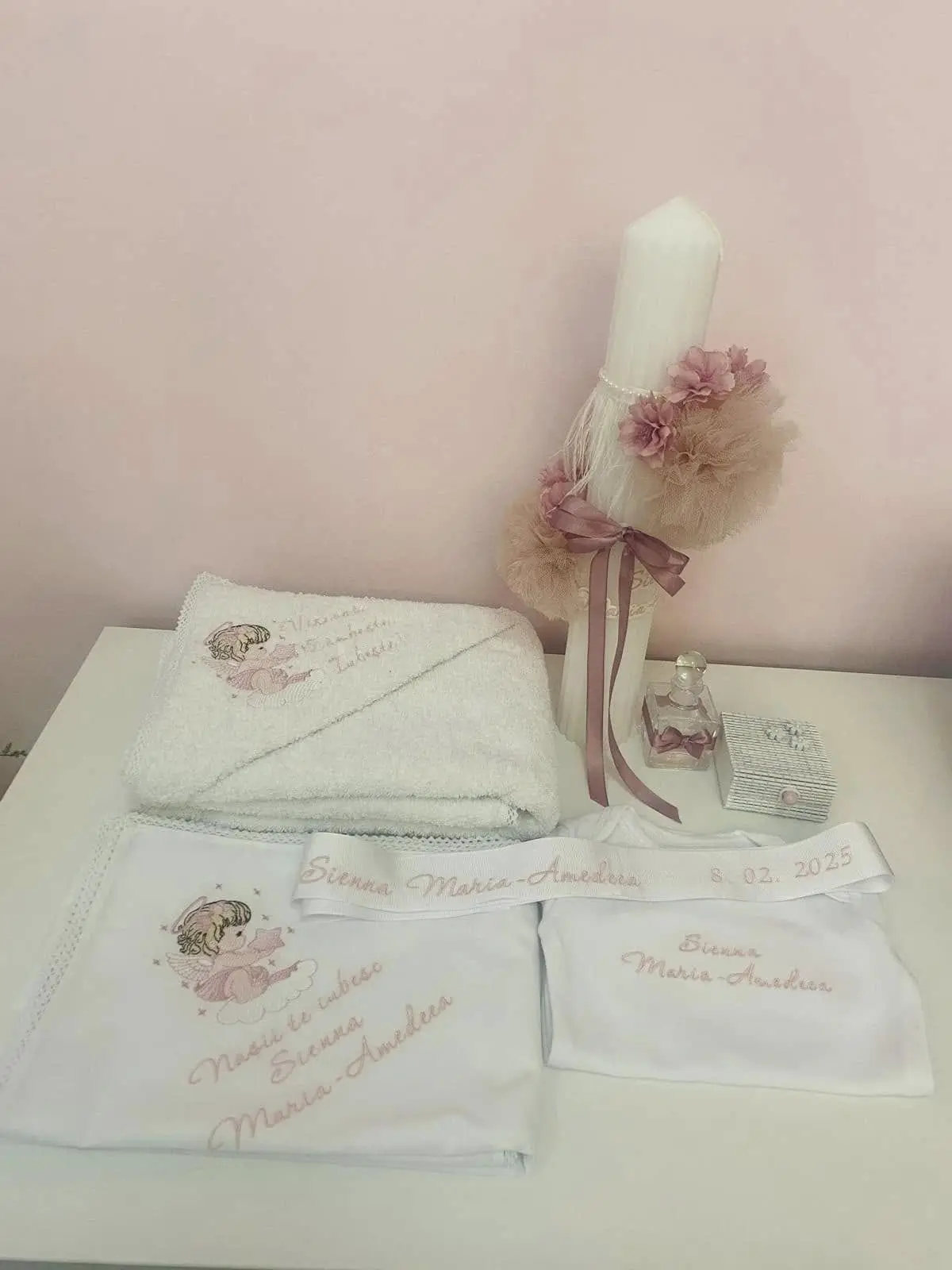 Trusou de botez cu prosoape brodate cu îngeraș și numele fetiței, lângă lumânare cu flori roz